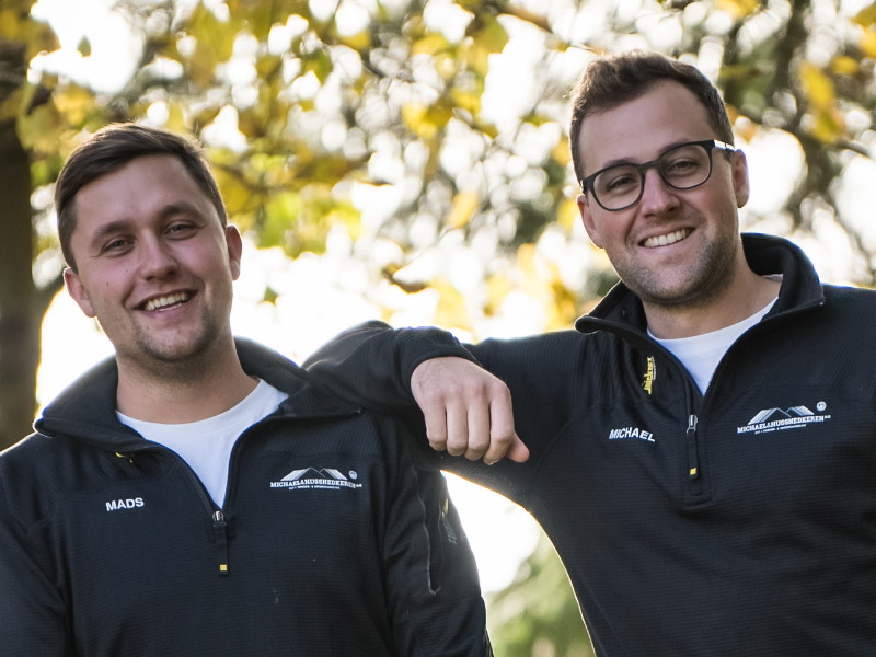 To smilende mænd, Mads og Michael, i sorte arbejdsjakker udendørs.